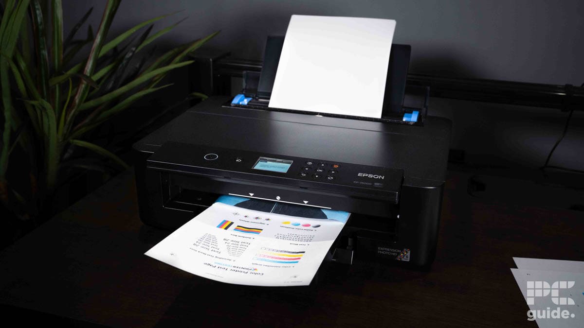 An Epson Expression XP-15000 printer on a desk ejecting a high-quality printed page with color charts, next to a potted plant, under ambient lighting.