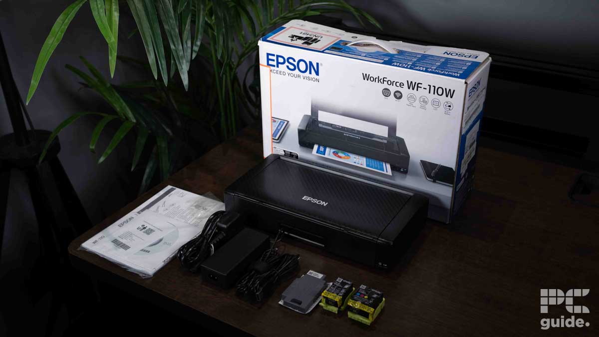 Epson WorkForce WF-110W compact printer box and printer on a table with accessories, including power cords, ink cartridges, manuals, and a CD.