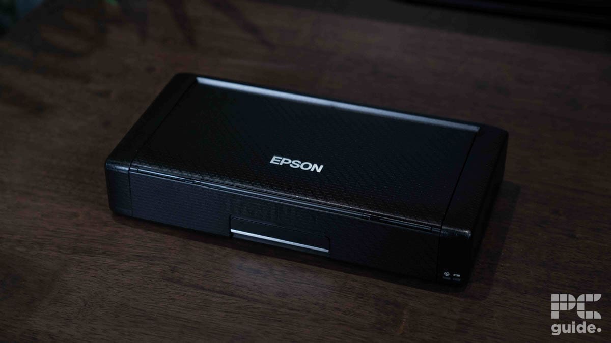 A closed black Epson WorkForce WF-110 portable printer sits on a wooden surface, with the brand name centered on the top. Compact and light, the device features the PCWer logo in the bottom right corner.